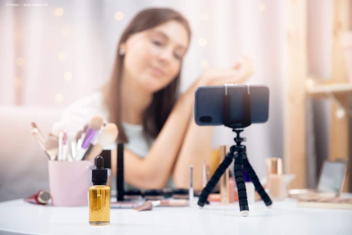 An influencer sitting in front of makeup and her phone with a vial of oil sitting on the table. (Image Credit: AdobeStock/Parilov)