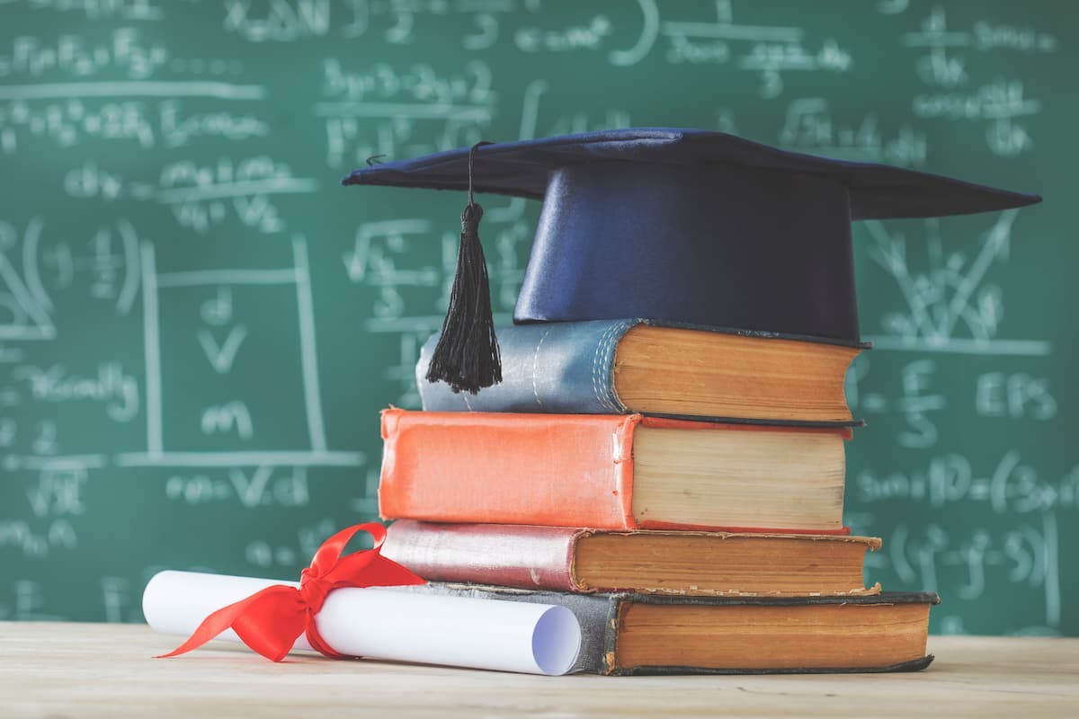 a stack of books in a classroom with a graduation cap and diploma sitting beside them. (Image Credit: AdobeStock/sebra)