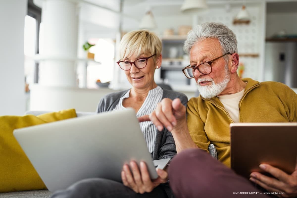 a senior couple sitting on the couch laughing and looking a laptop. (Image Credit: AdobeStock/NDABCREATIVITY)