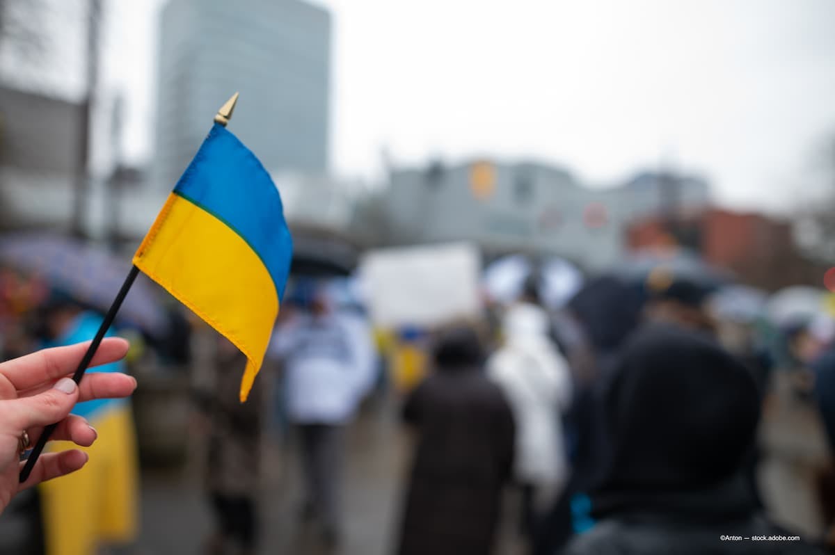 A hand is seen to the left of the frame holding a small Ukrainian flag.