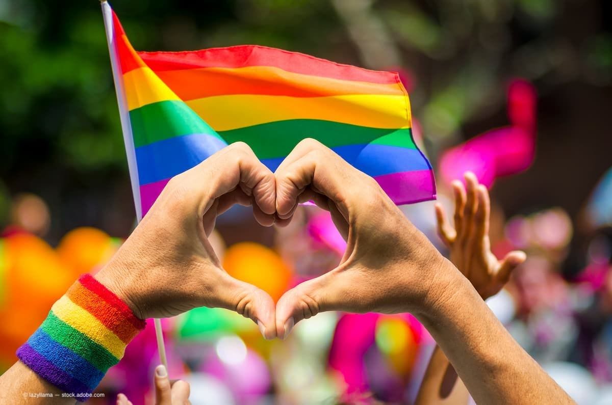 Hands making heart sign in front of rainbow flag. (Image Credit: AdobeStock/lazyllama)
