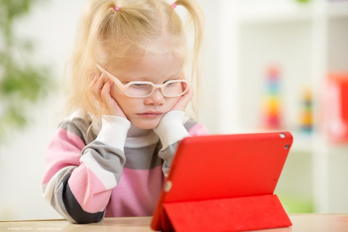 A young blonde girl with glasses sitting and staring at a red ipad. (Image Credit: AdobeStock/Andrey Kuzmin)