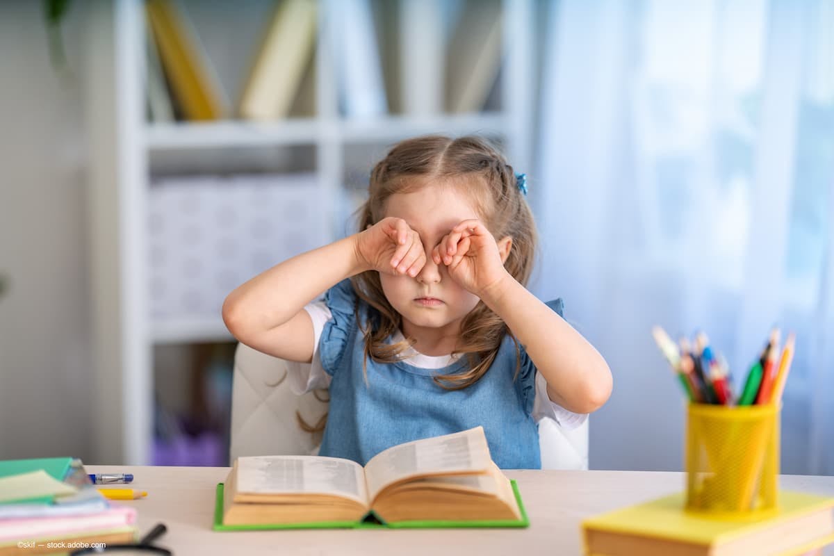 a child rubbing her eyes while reading a book. (Image Credit: AdobeStock/skif)